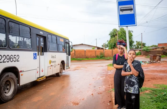 Duas crianças em uma rua de terra em um ponto de onibus esperando o onibus; Onibus ao fundo.